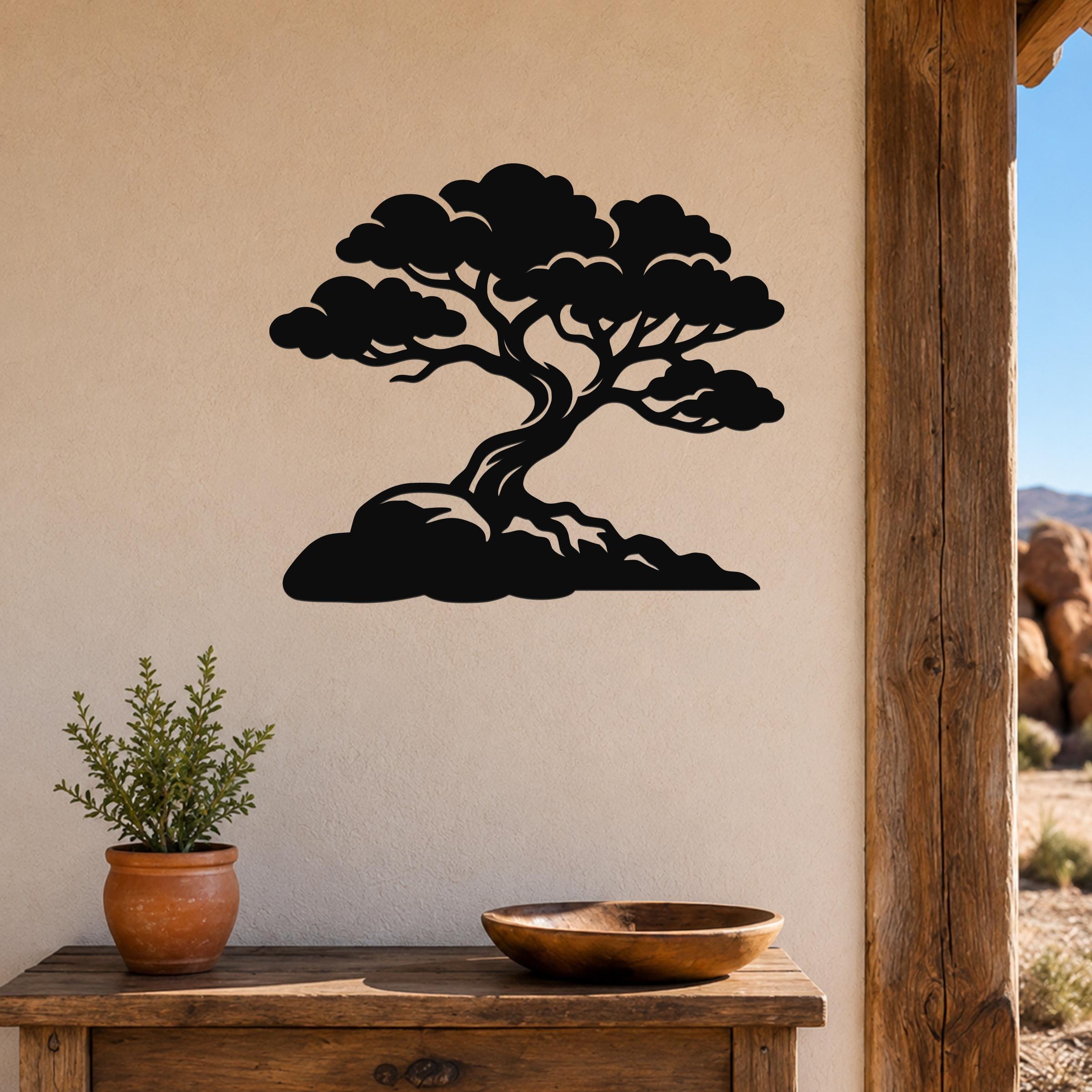 Black metal wall art cutout of a bonsai-style juniper tree with layered branches and roots, mounted on a beige wall above a wooden table in a rustic outdoor entryway.