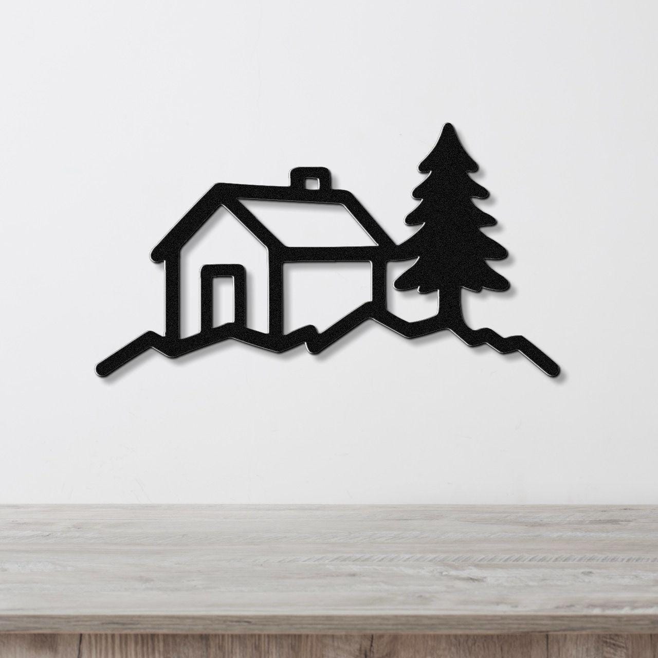 Black metal wall art cutout of a cabin and pine tree silhouette, mounted on a white wall above a light wood counter.