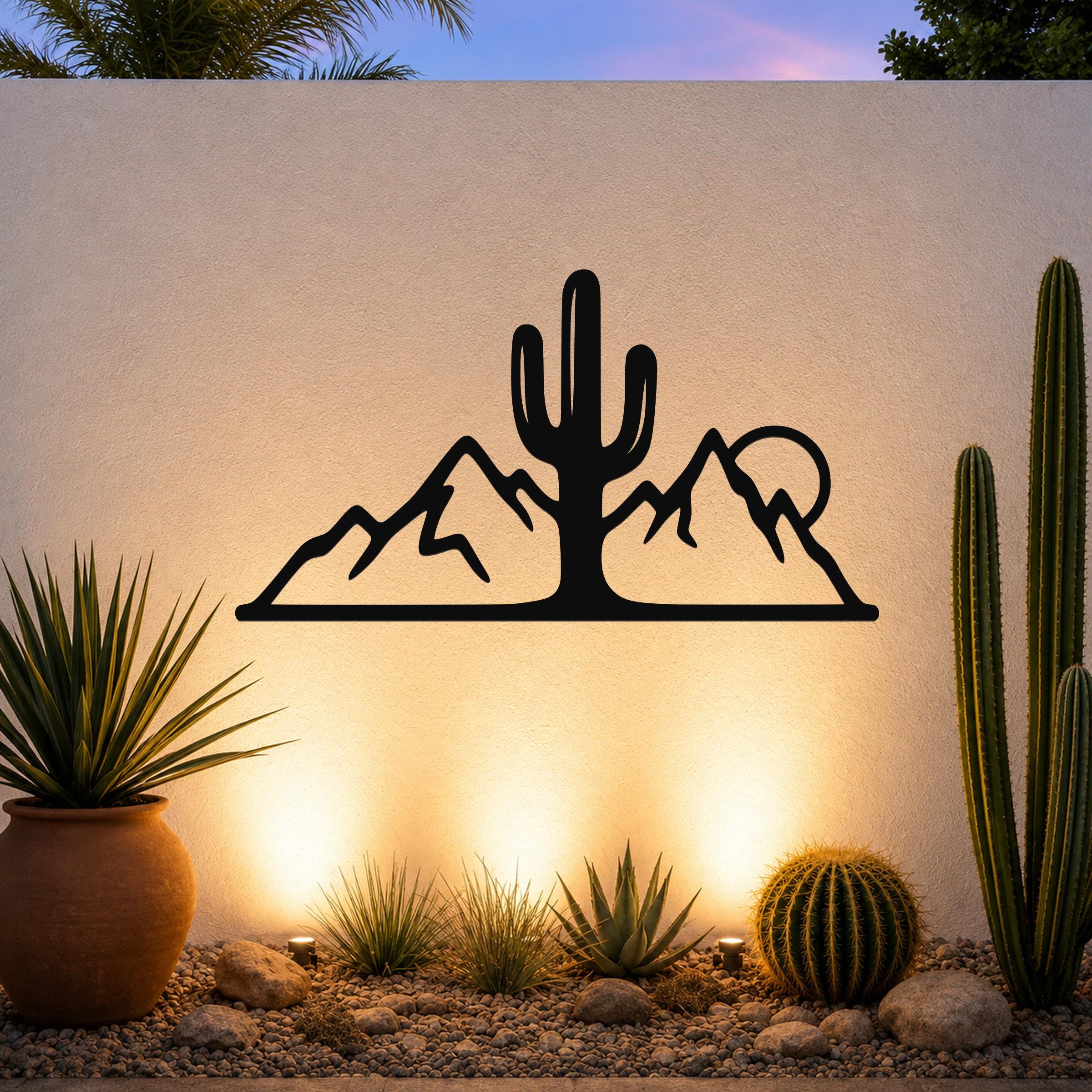 Black metal wall art with a saguaro cactus and mountain silhouette, mounted on an outdoor wall above a desert garden with potted plants and ground lights.