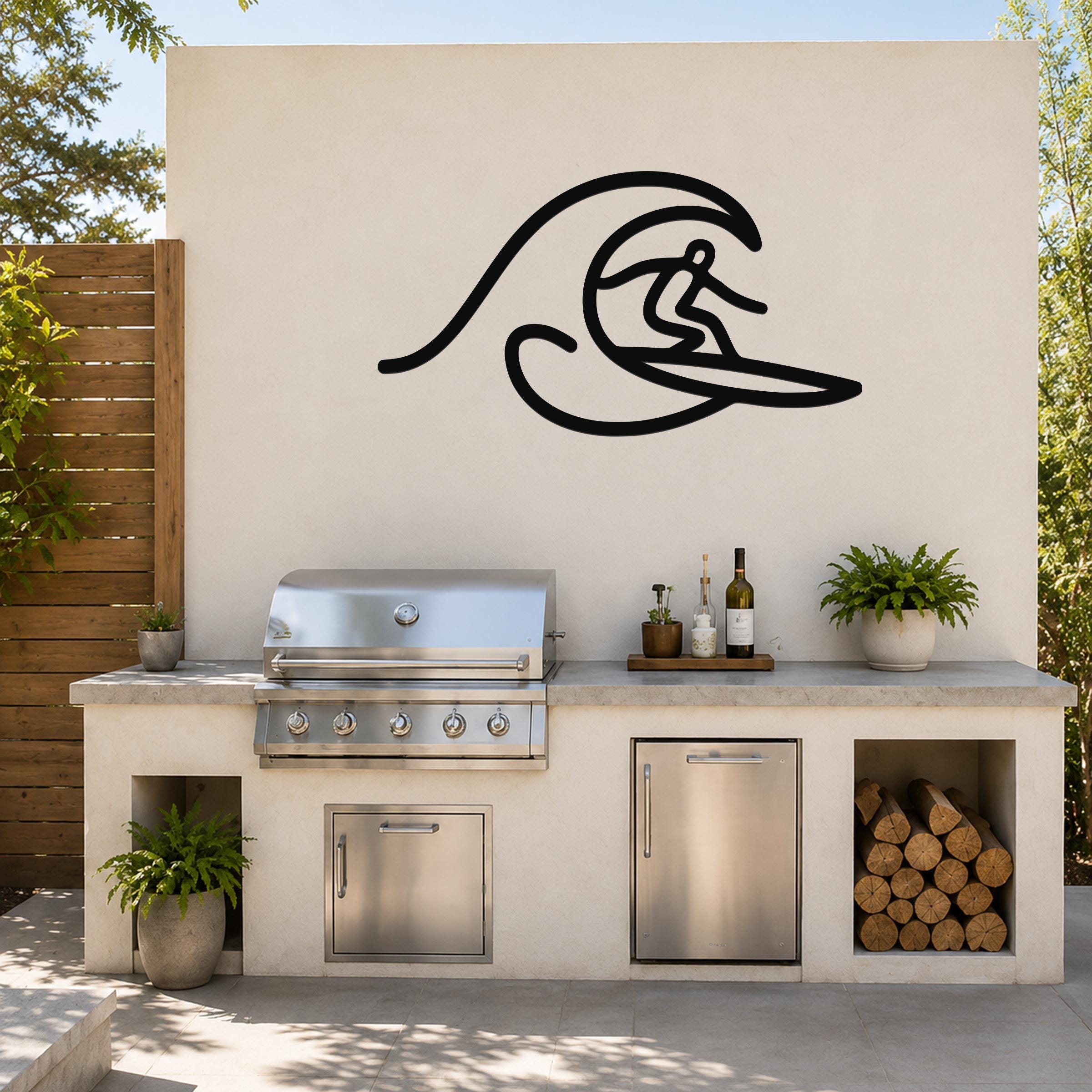 Black metal wall art silhouette of a surfer riding a wave, mounted on an outdoor kitchen wall above a stainless steel grill and counter.