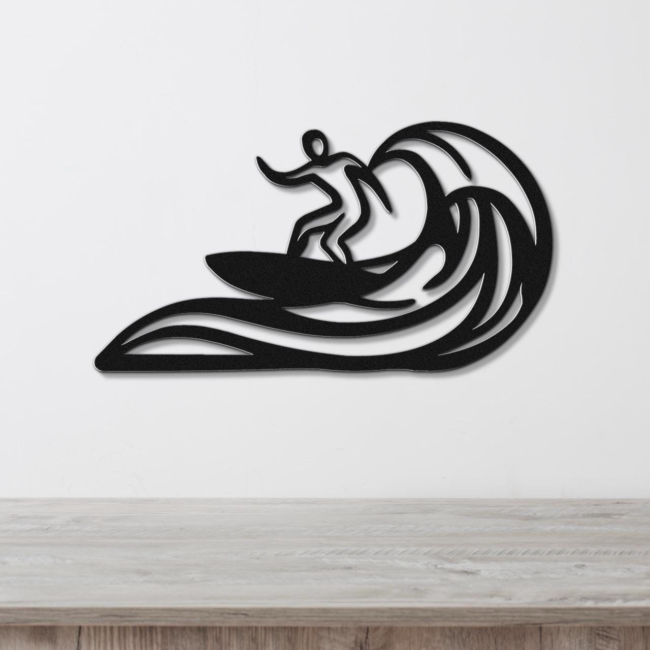 Black metal wall art featuring a surfer silhouette riding a wave, mounted on a white wall above a wooden surface.