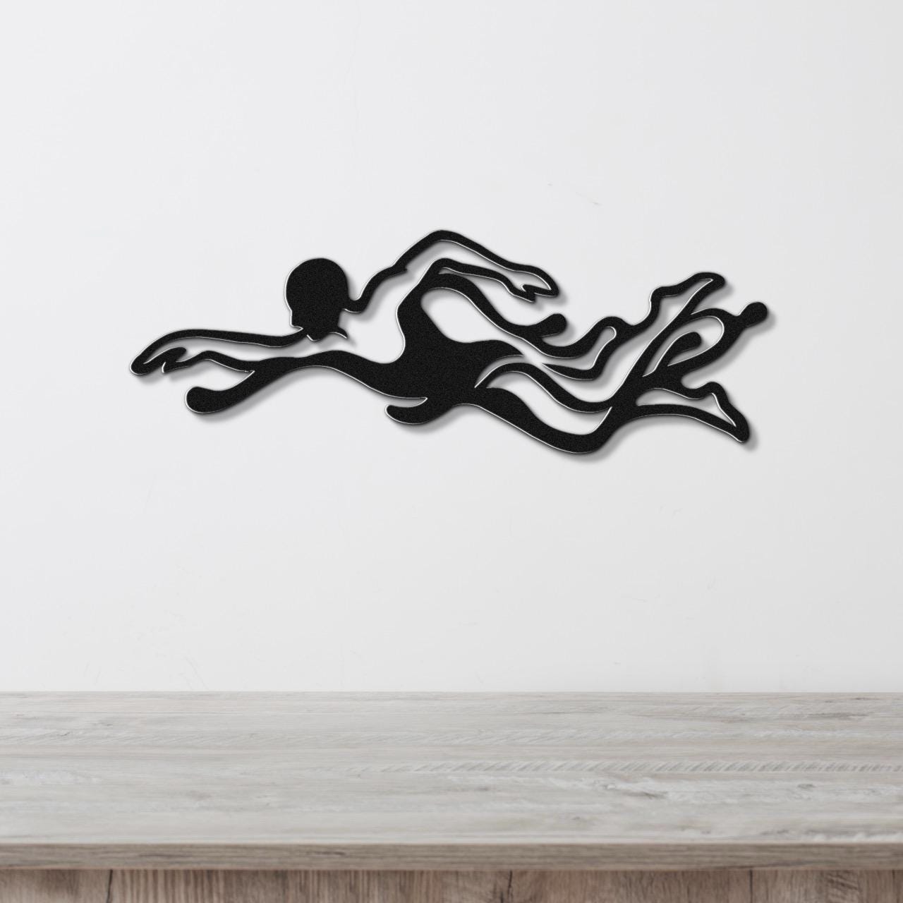 Black metal wall art of a swimmer in motion with flowing water cutouts, mounted on a white wall above a light wood surface.