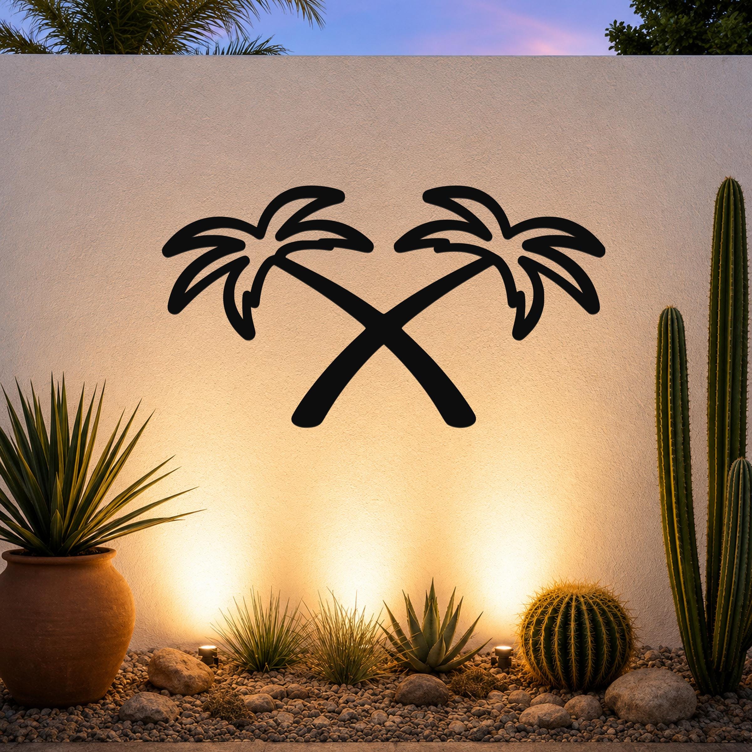 Black metal wall art featuring two crossed palm trees in silhouette, mounted on an outdoor wall above a desert garden with cacti and warm uplighting.