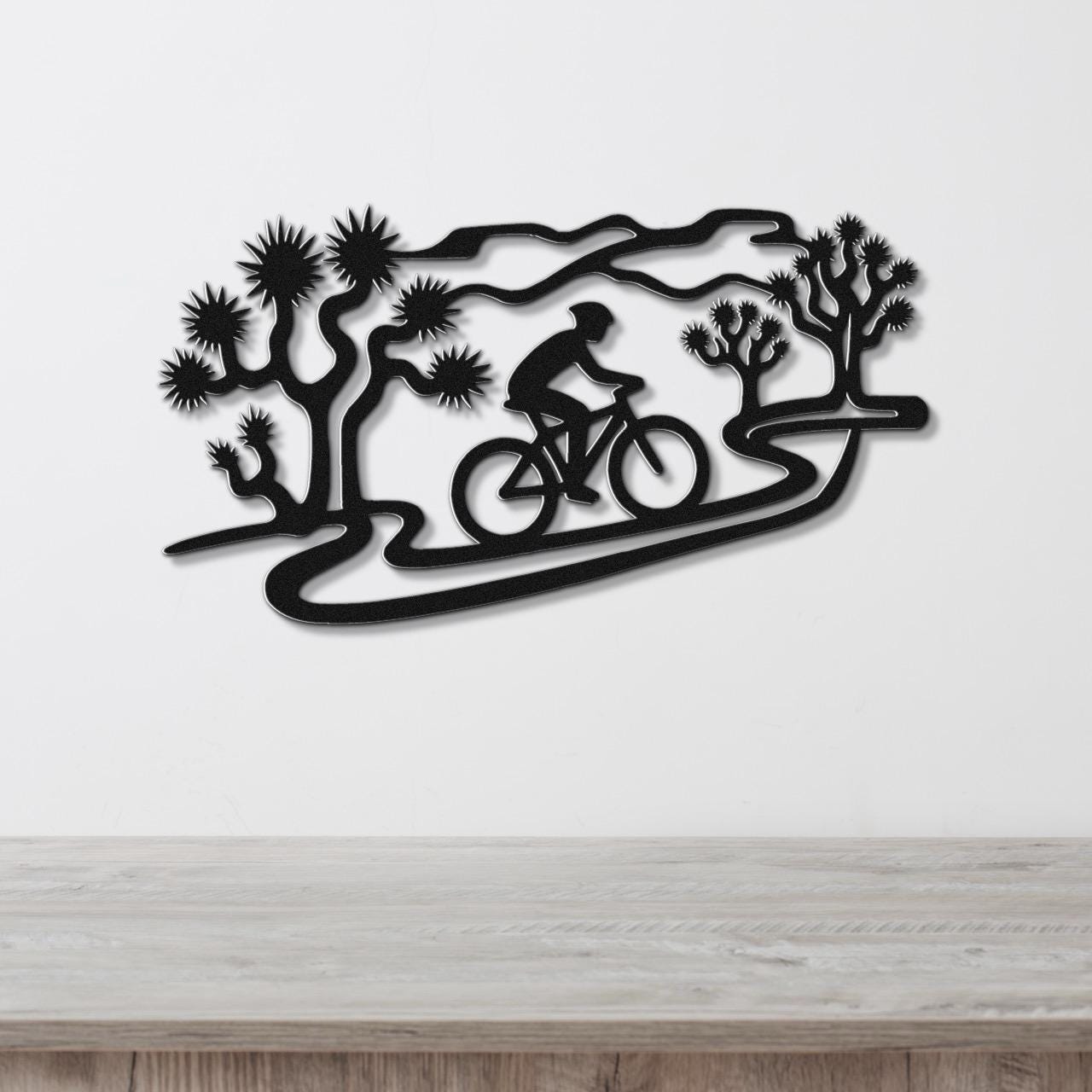 Black metal wall art featuring a cyclist silhouette on a winding path surrounded by Joshua trees, mounted on a white wall above a wooden surface.