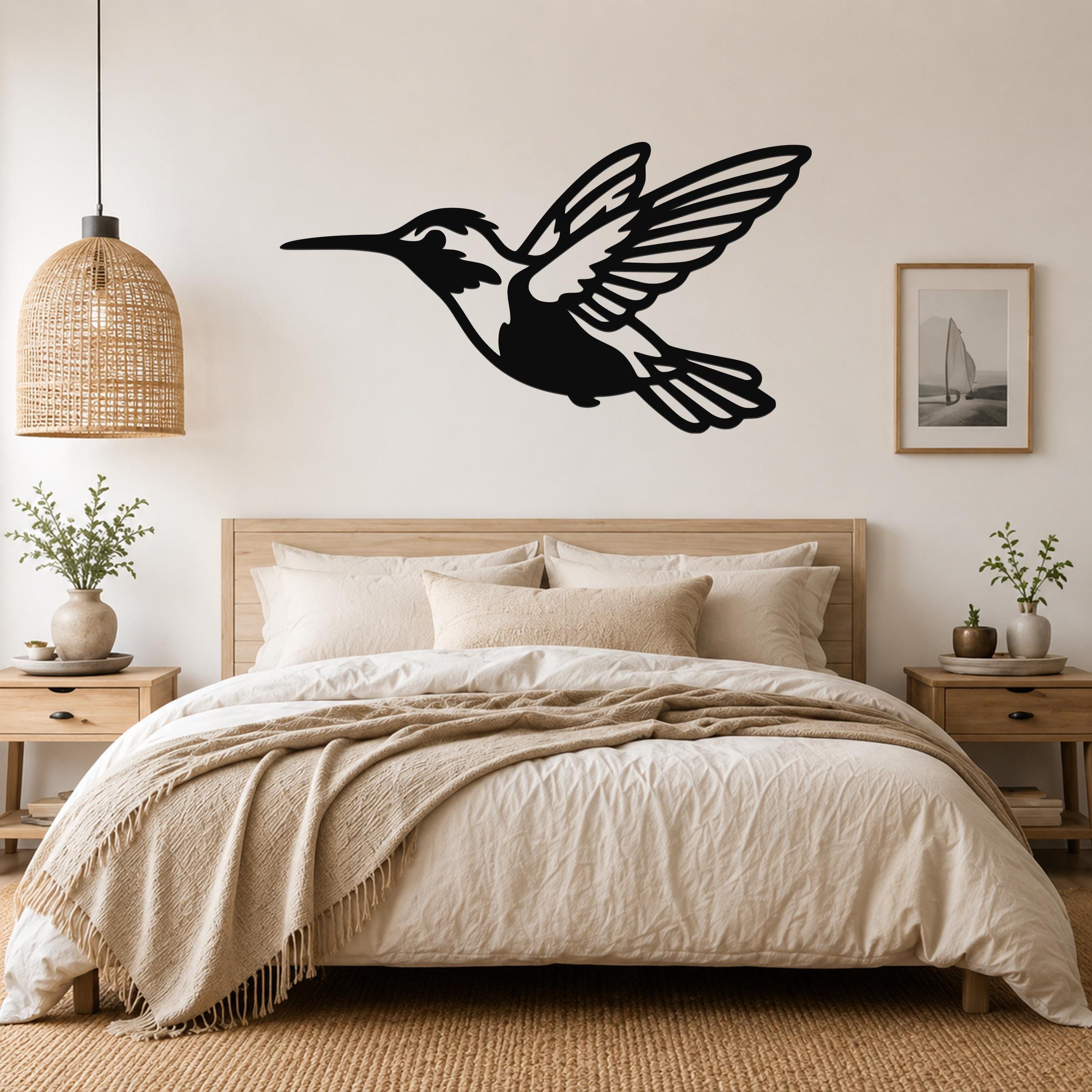 Black metal hummingbird wall art with open wings and internal cutout details, mounted above a wooden bed in a neutral bedroom.