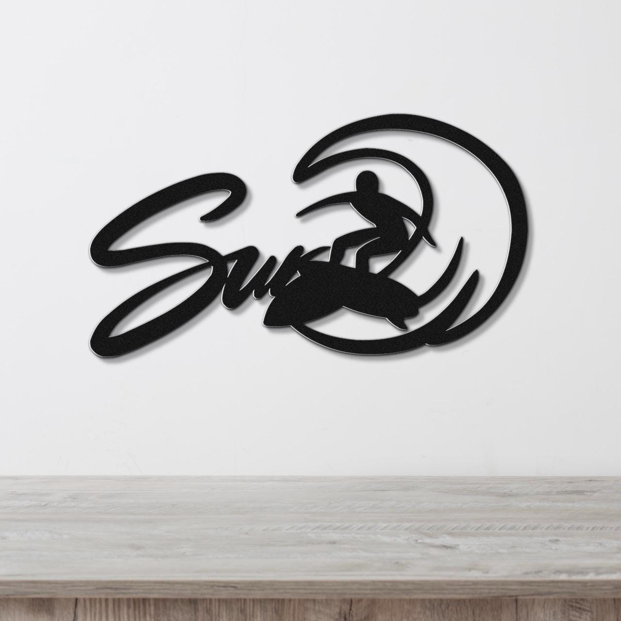 Black metal wall art featuring a surfer silhouette riding a wave, with the word "Surf" in cursive script, mounted on a white wall above a light wood surface.