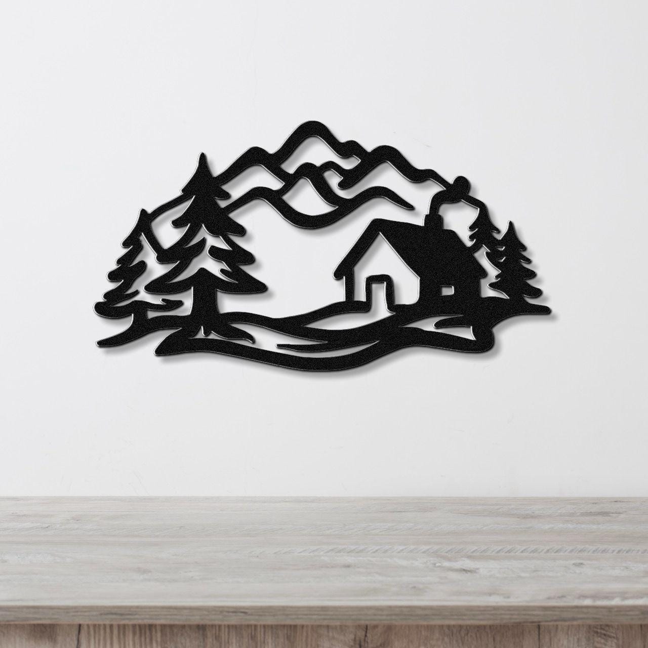 Black metal wall art featuring a mountain cabin silhouette with pine trees and layered peaks, mounted on a white wall above a wooden surface.