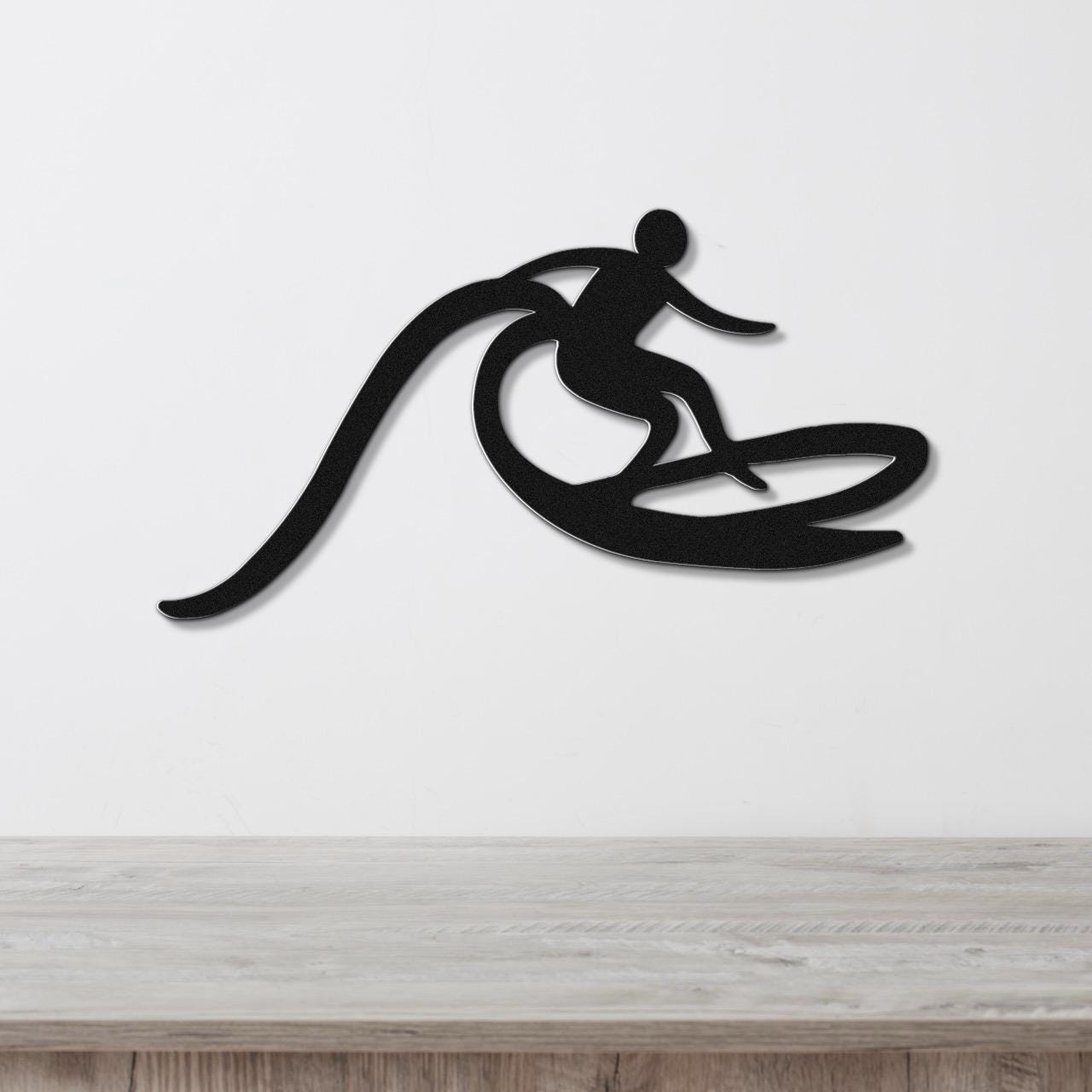Black metal wall art cutout of a surfer riding a wave, mounted on a white wall above a light wood counter.