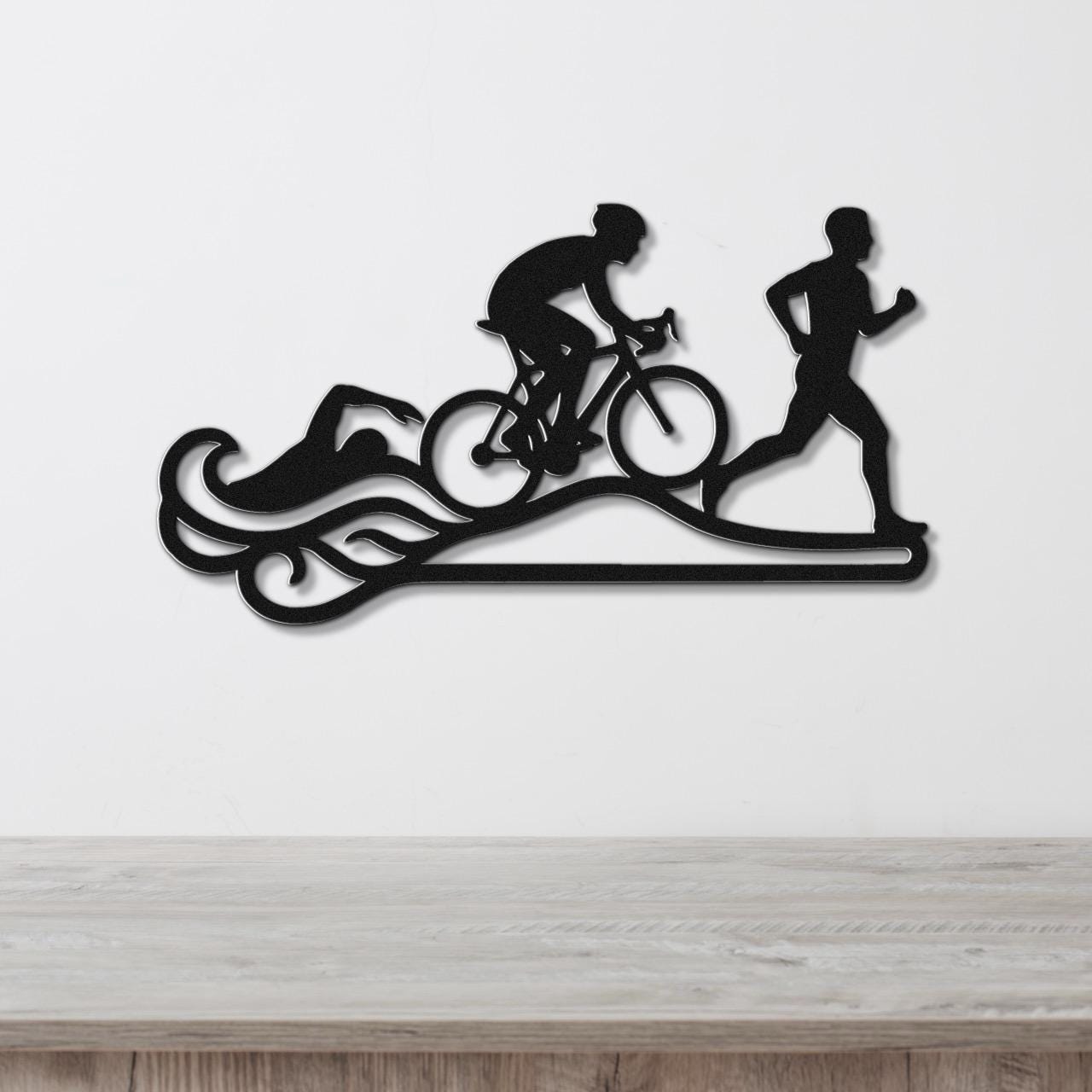 Black metal triathlon wall art with swim, bike, and run silhouettes and motion lines, mounted on a white wall above a wooden surface.