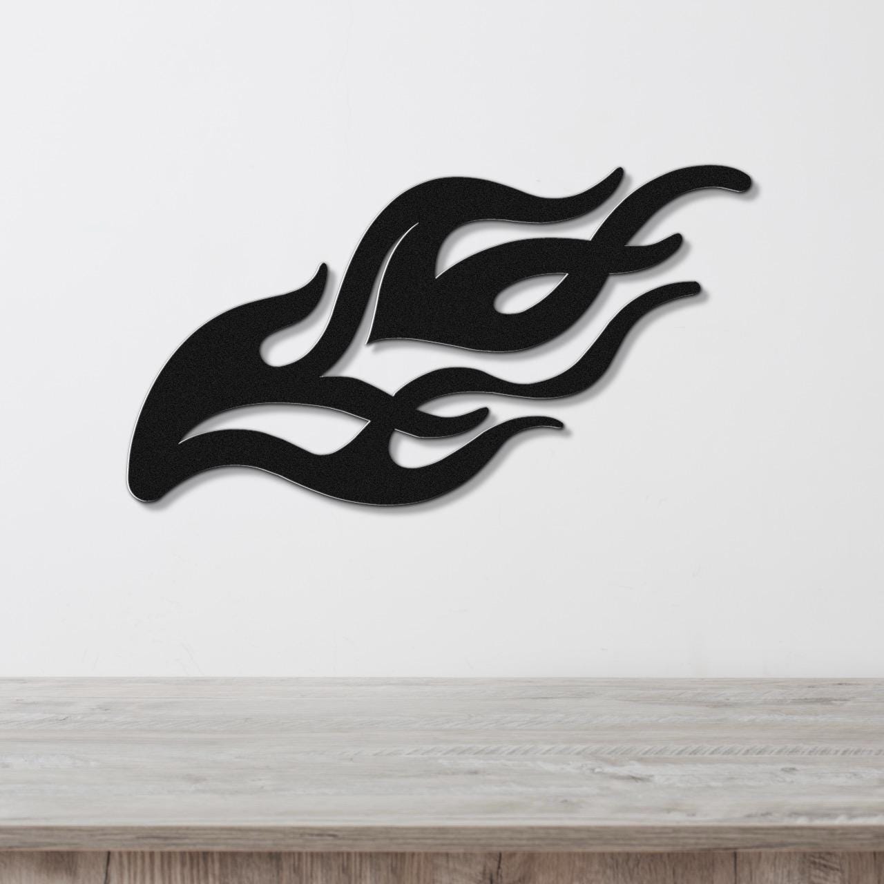 Black metal flame wall art with flowing silhouette cutouts, mounted on a white wall above a light wood surface.