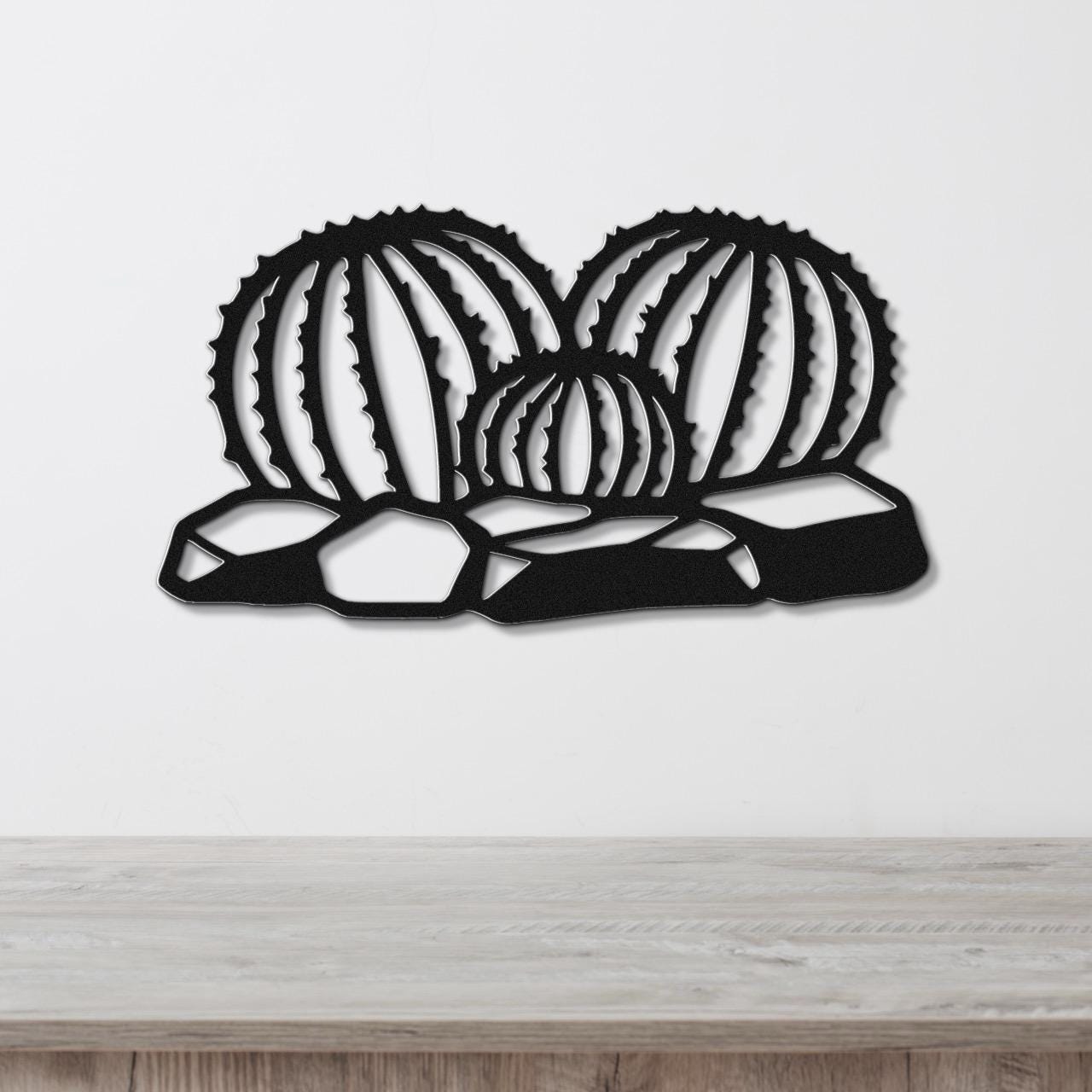 Black metal barrel cactus wall art with ribbed cutout design, mounted on a white wall above a wooden surface.