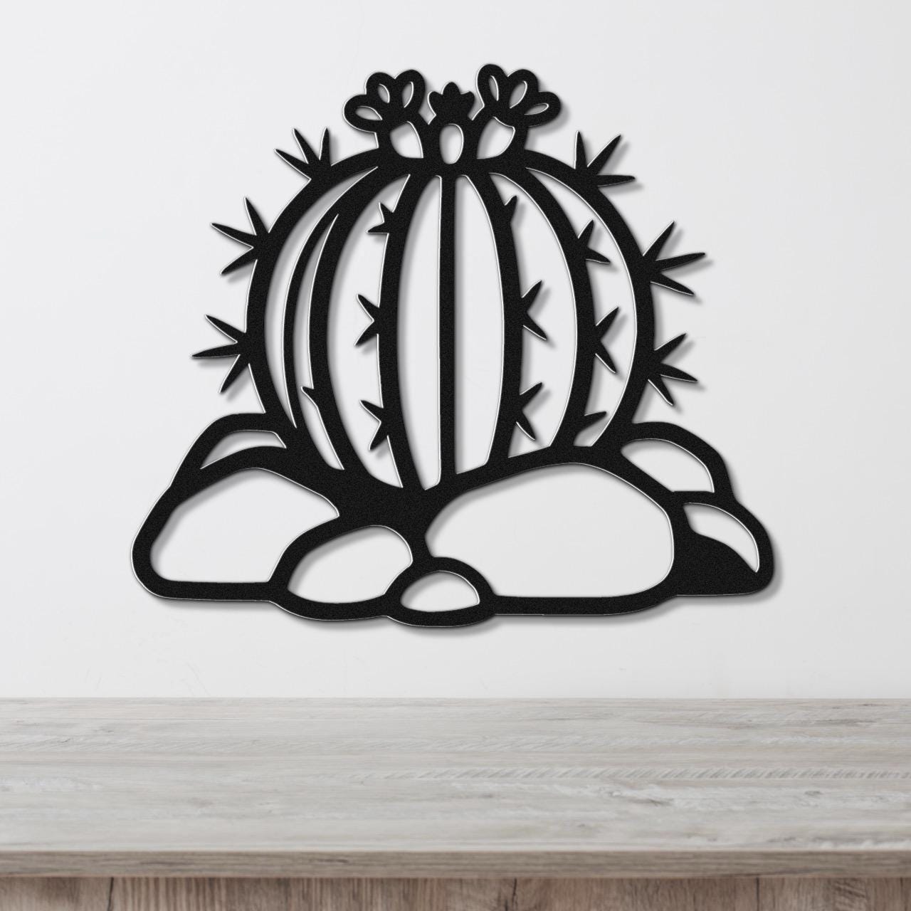 Black metal barrel cactus wall art with flower and spine cutouts, mounted on a white wall above a wooden counter.