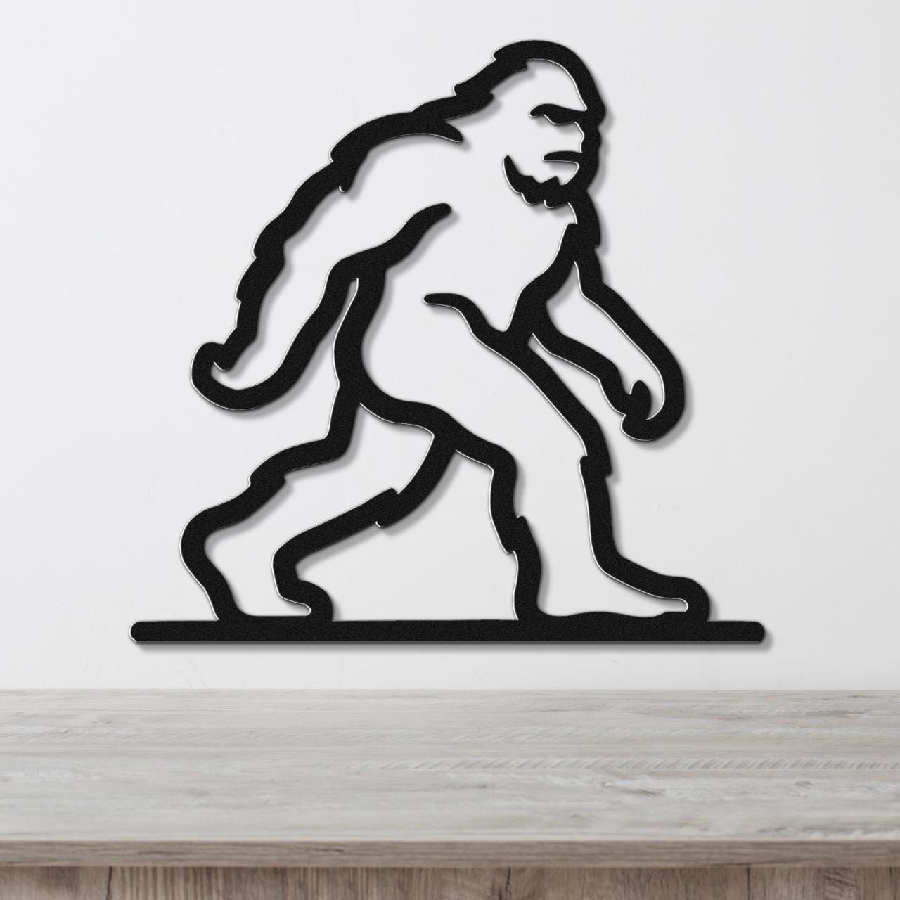 Black metal wall art silhouette of Bigfoot in a walking pose, mounted on a white wall above a wooden surface.