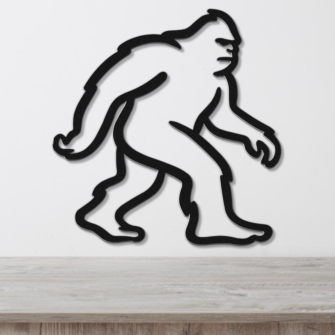 Black metal wall art featuring a minimalist Bigfoot silhouette, mounted on a white wall above a wooden surface.