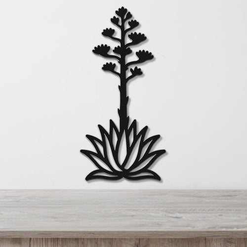 Black metal wall art in the silhouette of a blooming agave plant with a tall flower stalk and spiky rosette base, mounted on a white wall above a light wood surface.