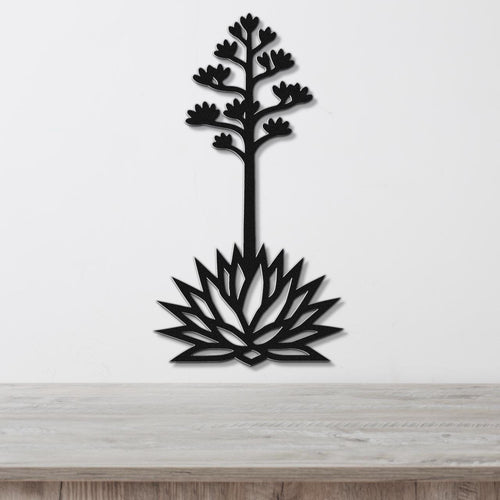 Black metal wall art cutout of a blooming agave plant with a tall stem and flower spikes, mounted on a white wall above a light wood surface.