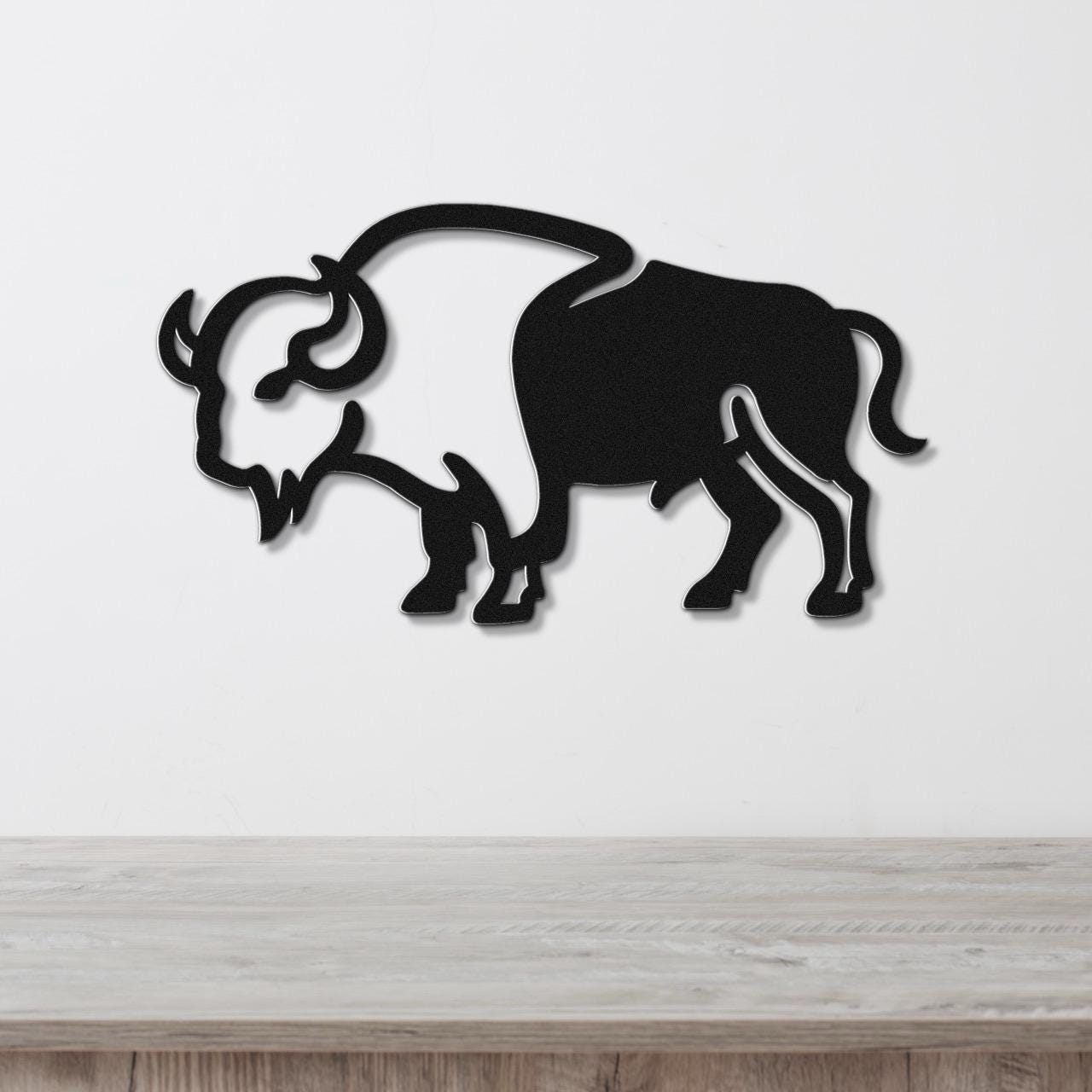 Black metal buffalo wall art silhouette with cutout horns and mane, mounted on a white wall above a light wood surface.