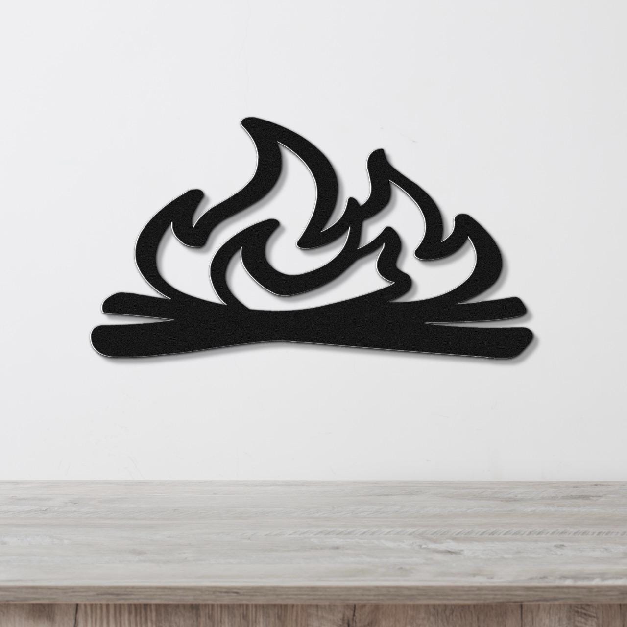 Black metal wall art with a flame and log silhouette, mounted on a white wall above a light wood surface.