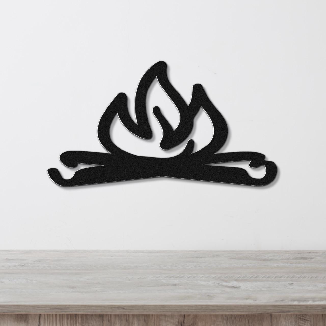 Black metal wall art cutout of a campfire with flames and logs, mounted on a white wall above a wooden surface.