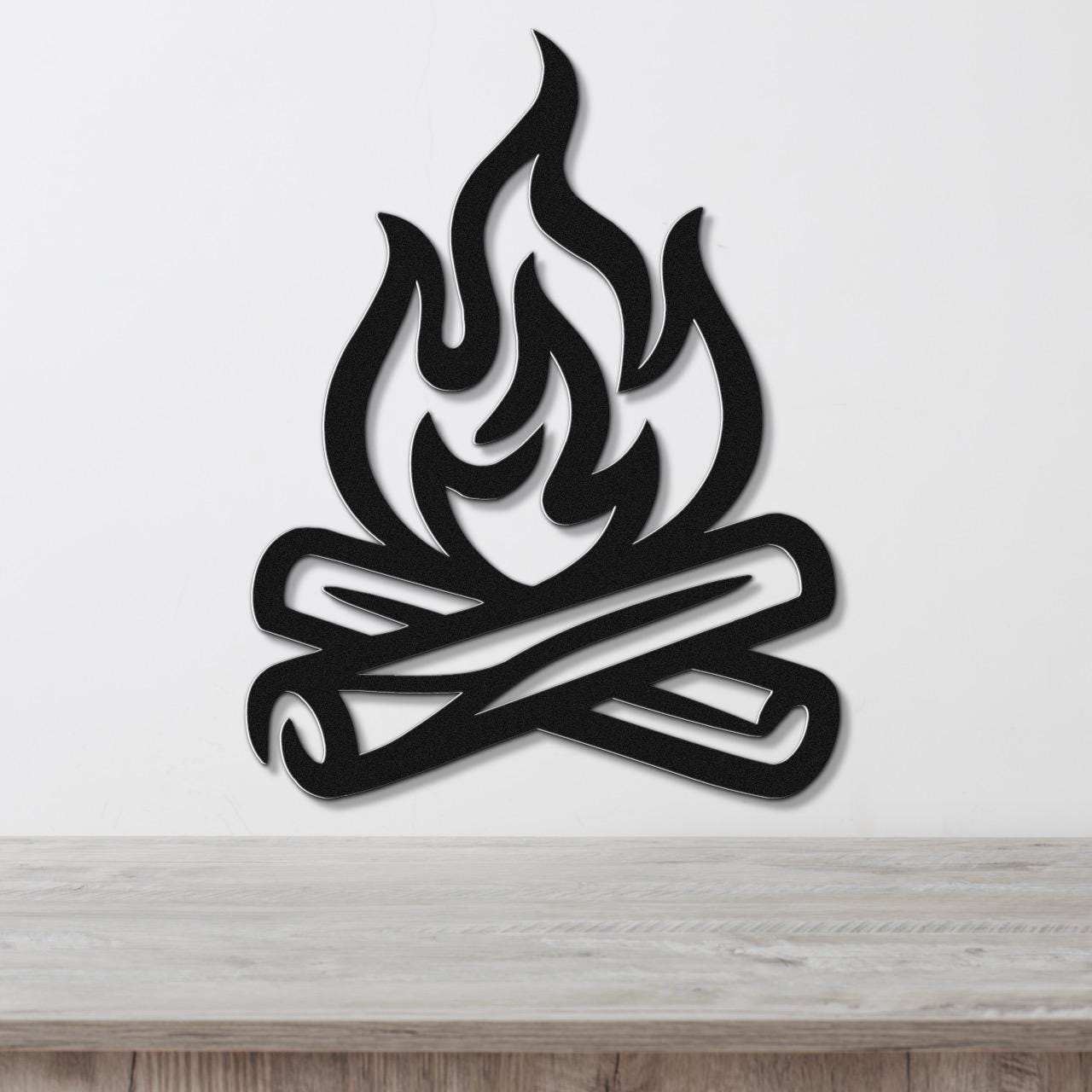 Black metal wall art in a campfire silhouette design with crossed logs and flame cutouts, mounted on a white wall above a wooden surface.