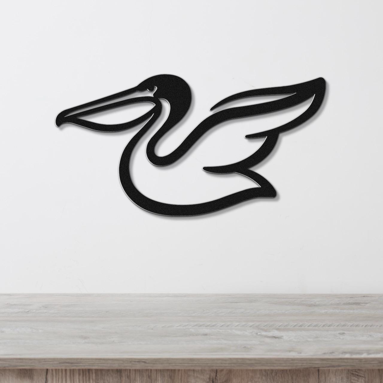 Black metal pelican wall art with a sleek silhouette and wing cutout, mounted on a white wall above a light wood surface.