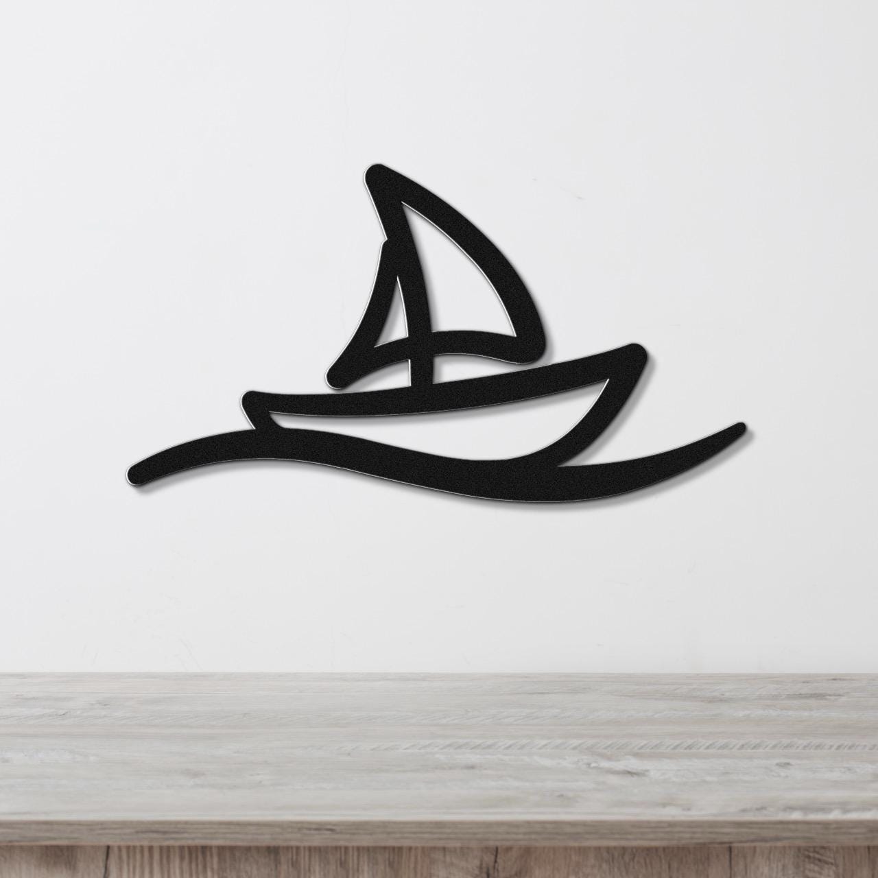 Black metal wall art in a sailboat silhouette with a cutout sail and wave design, mounted on a white wall above a wooden surface.