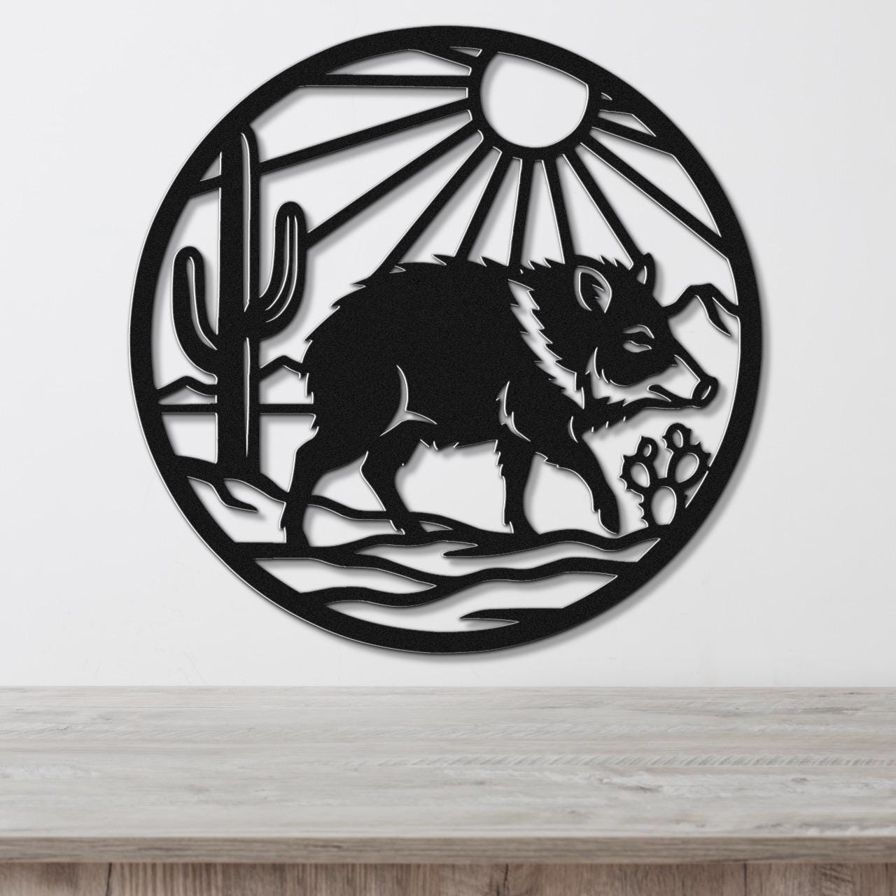 Black metal circular wall art featuring a javelina silhouette, cactus, sun rays, and paw prints, mounted on a white wall above a wooden surface.