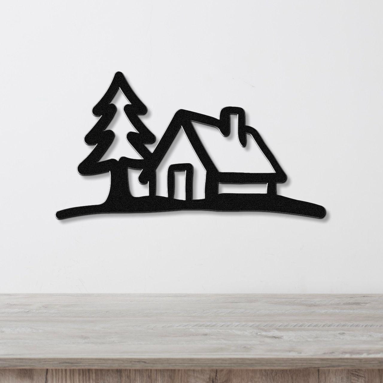 Black metal wall art cutout of a cabin and pine tree silhouette, mounted on a white wall above a wooden surface.