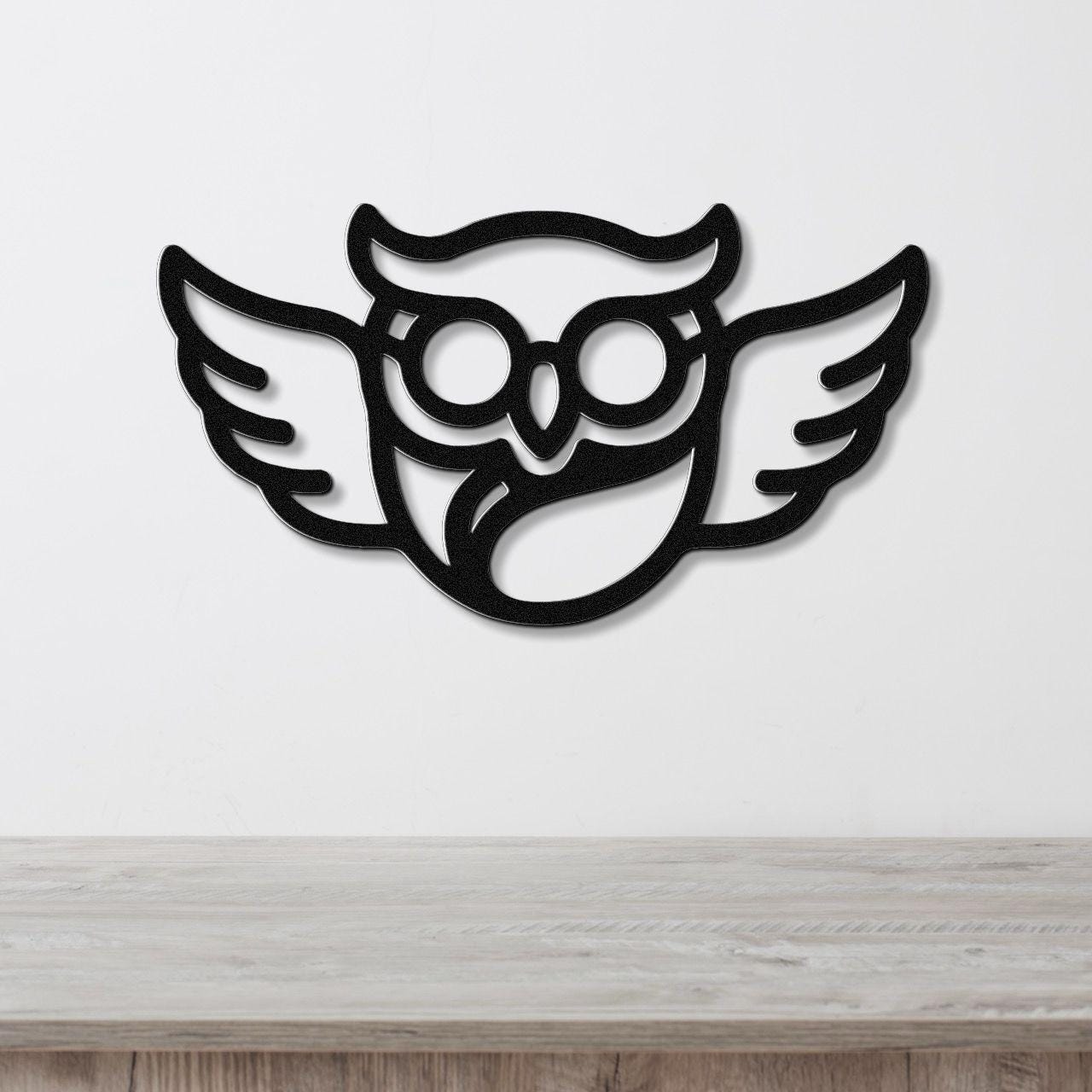 Black metal wall art featuring a bespectacled owl silhouette with wings, mounted on a white wall above a wooden surface.