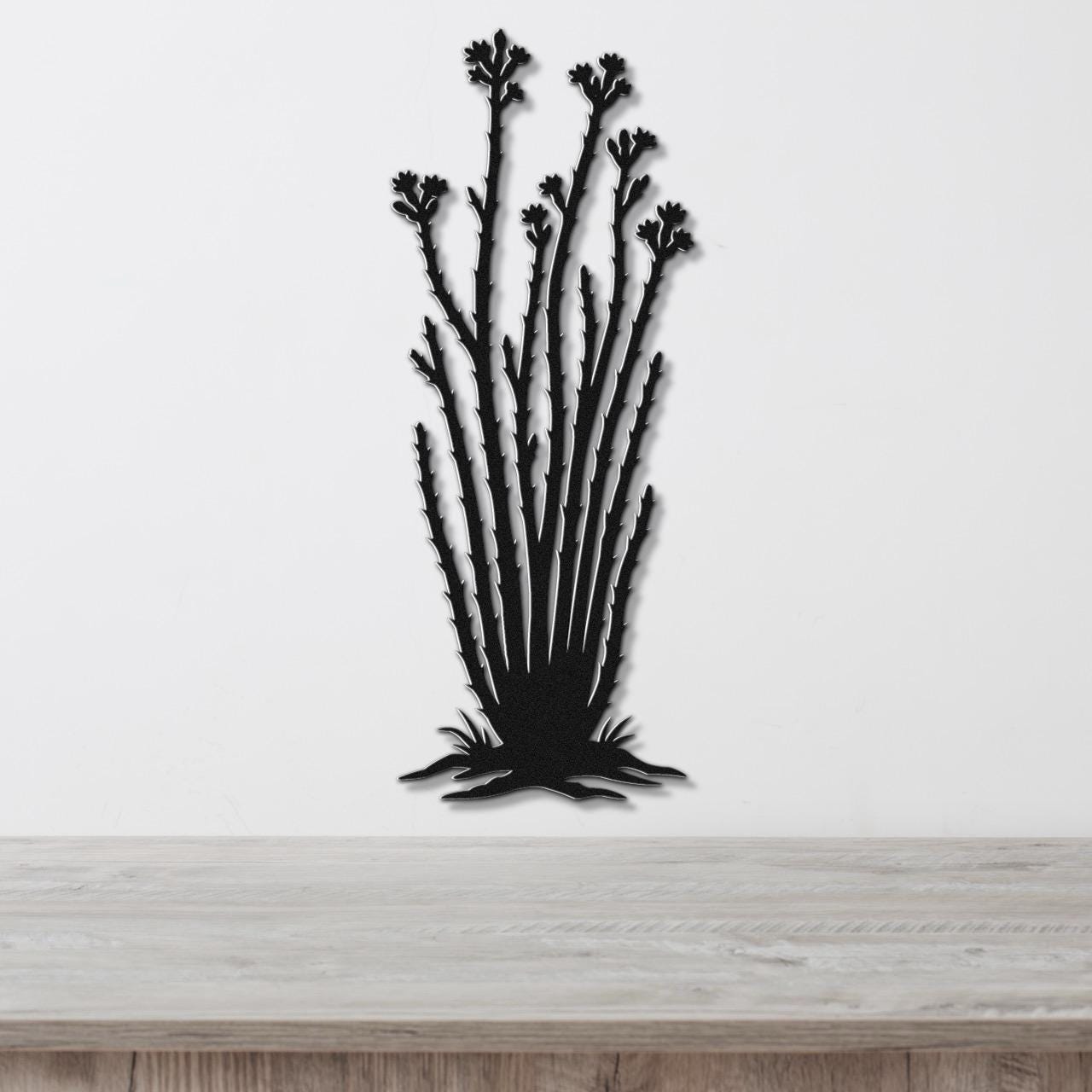 Black metal wall art cutout of an ocotillo plant with branching stems and flower clusters, mounted on a white wall above a light wood surface.
