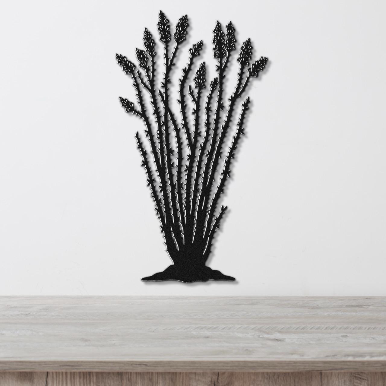 Black metal wall art silhouette of an ocotillo plant with spiny stems and clustered flower cutouts, mounted on a white wall above a light wood surface.