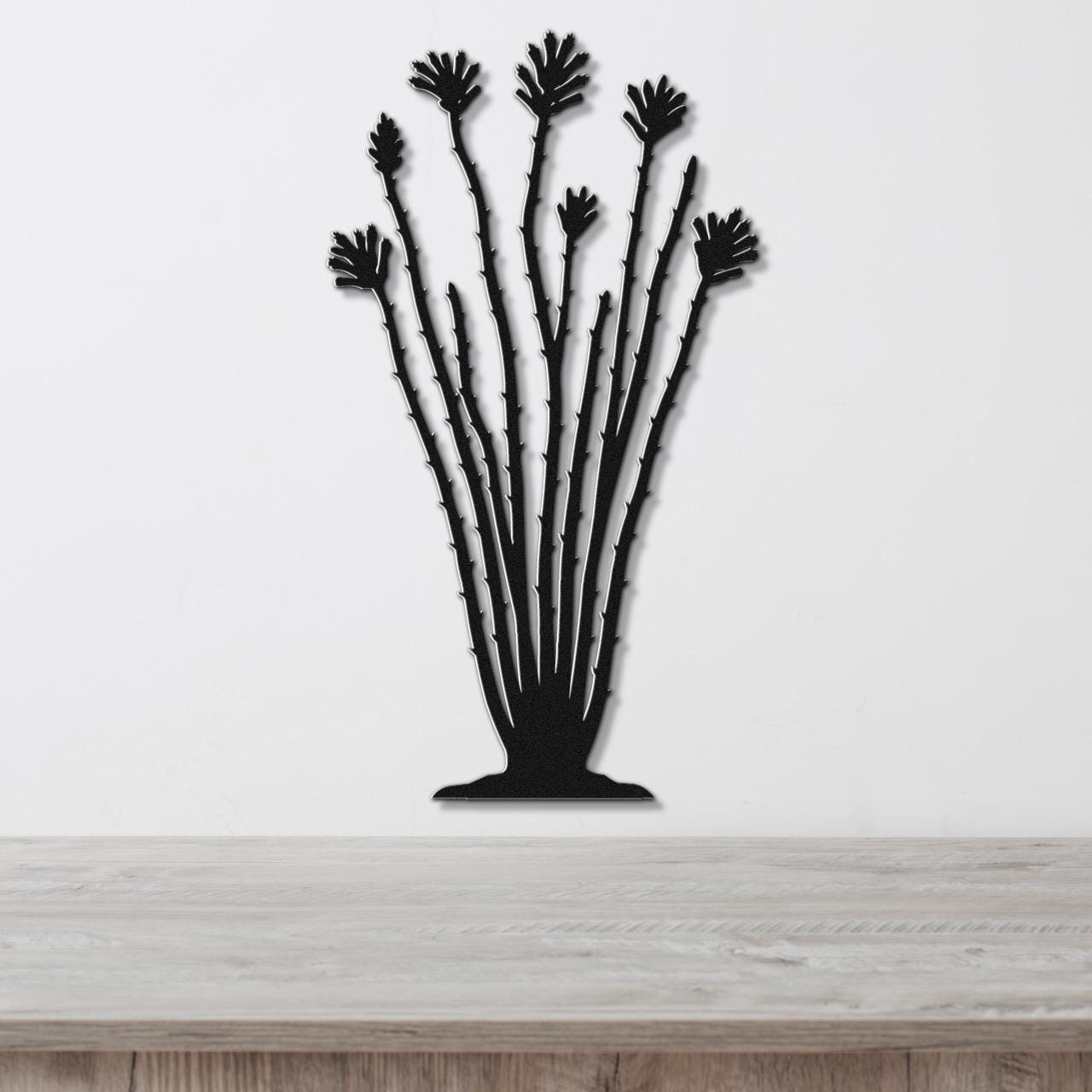 Black metal ocotillo wall sculpture with spiny stems and fan-shaped leaf cutouts, mounted on a white wall above a wooden surface.
