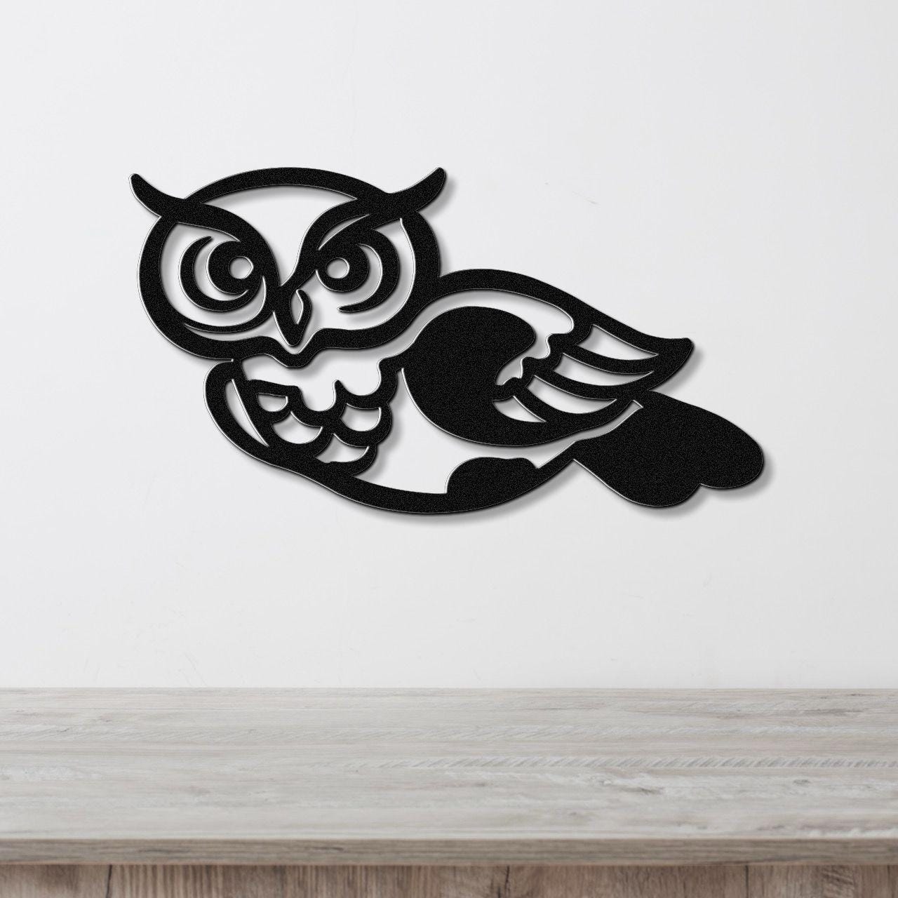 Black metal owl wall art with intricate cutout details of eyes, wings, and feathers, mounted on a white wall above a light wood surface.