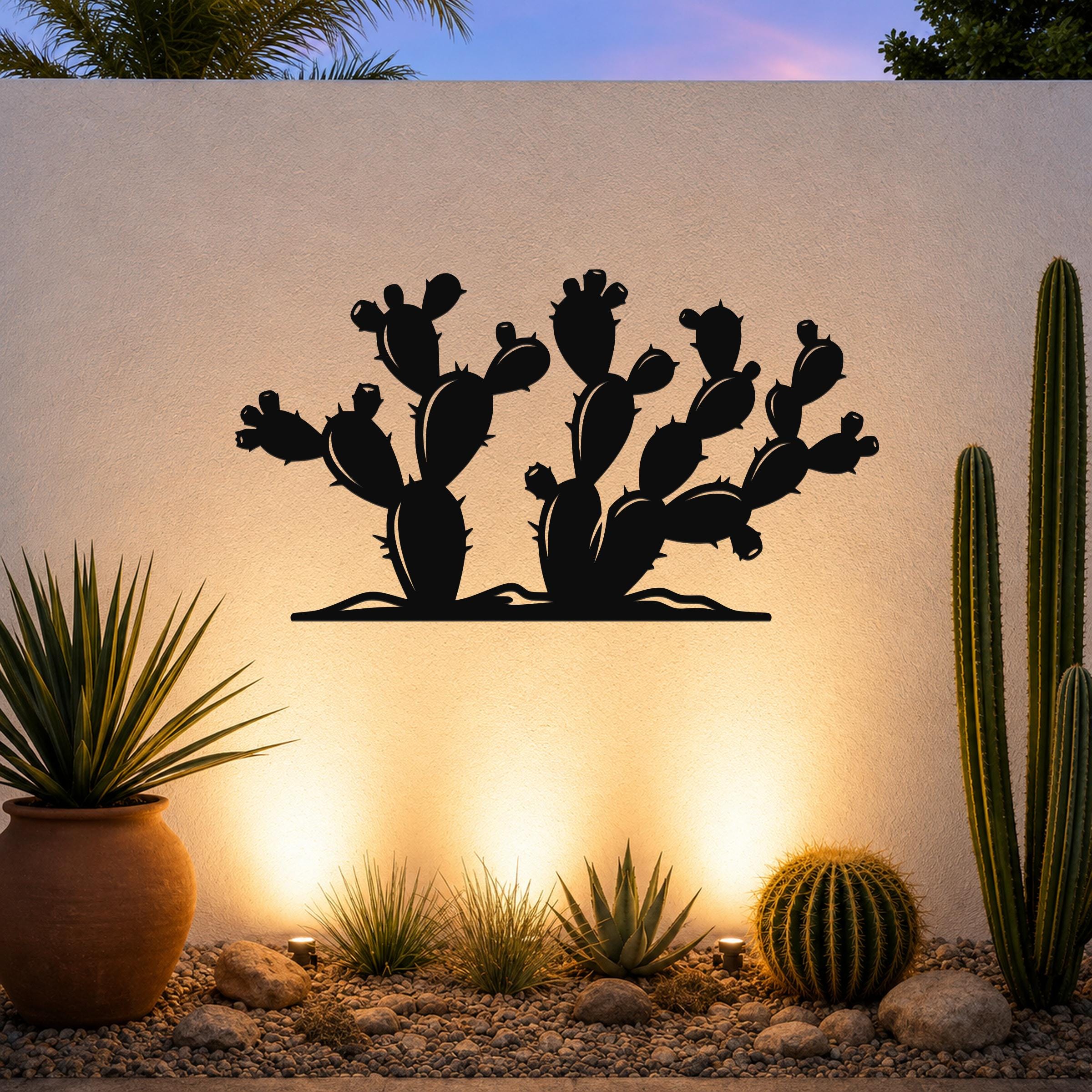 Black metal cactus wall art with prickly pear silhouette, mounted on a textured outdoor wall above a gravel garden with potted plants and uplighting.
