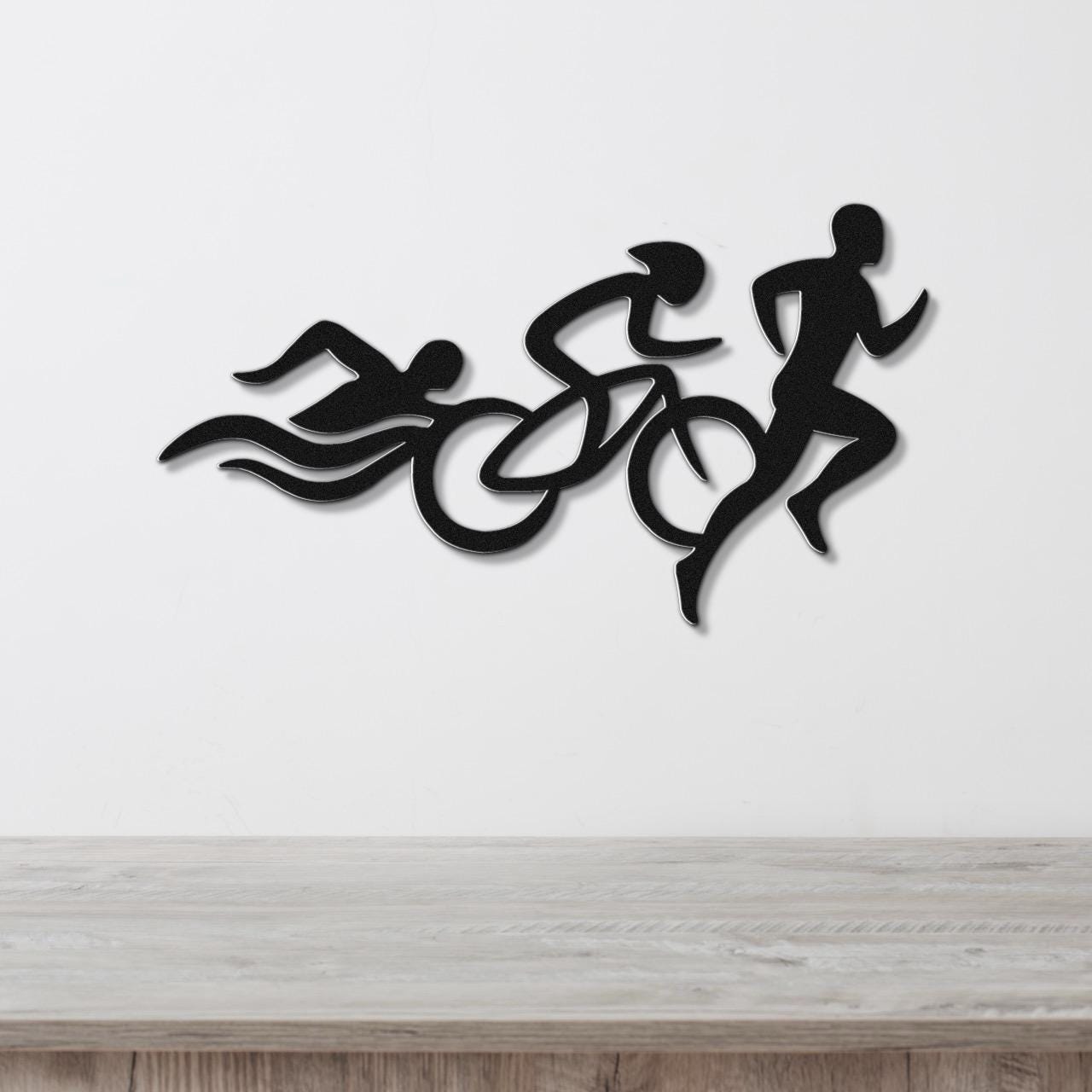 Black metal wall art featuring a triathlon silhouette of swim, bike, and run figures in motion, mounted on a white wall above a wooden surface.