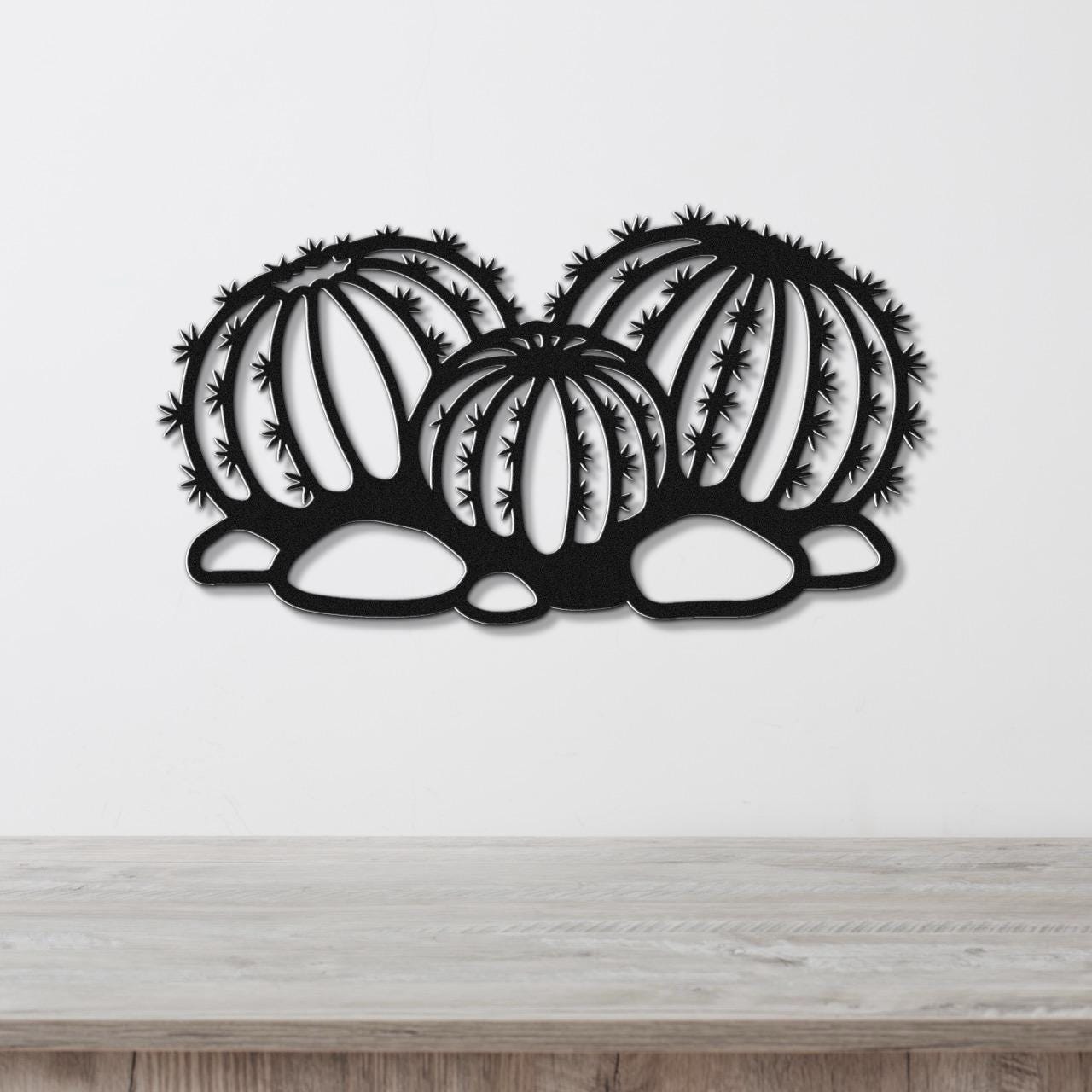 Black metal barrel cactus wall art with spines and vertical ribs cutout, mounted on a white wall above a wooden surface.