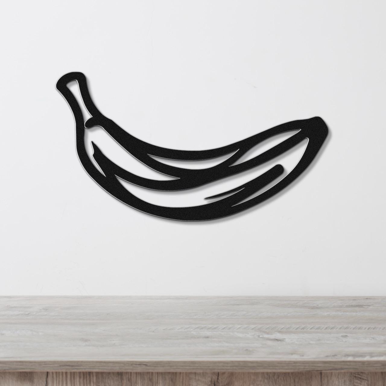 Black metal banana wall art with internal cutout lines, mounted on a white wall above a light wood countertop.