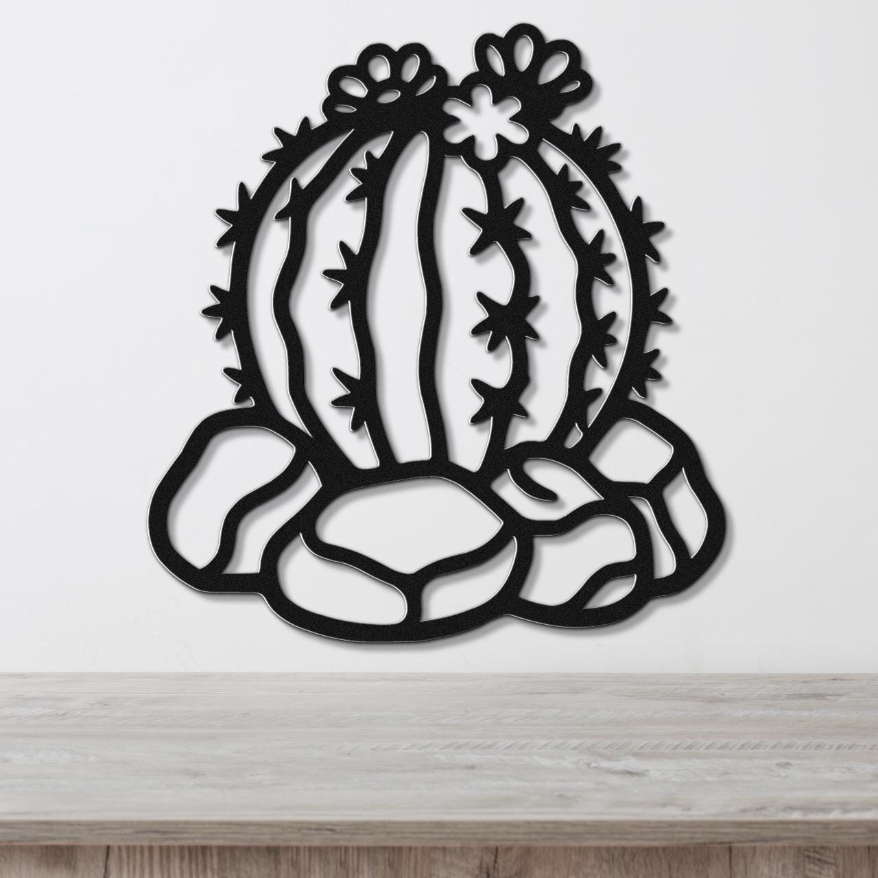 Black metal barrel cactus wall art with flower and spine cutouts, mounted on a white wall above a wooden surface.