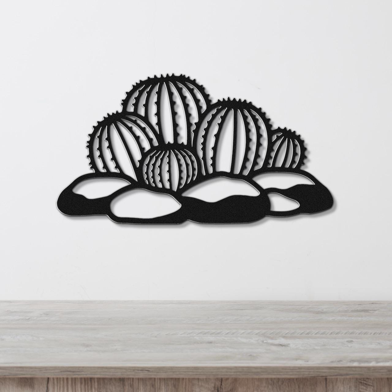 Black metal barrel cactus wall art with geometric cutouts, mounted on a white wall above a wooden surface.