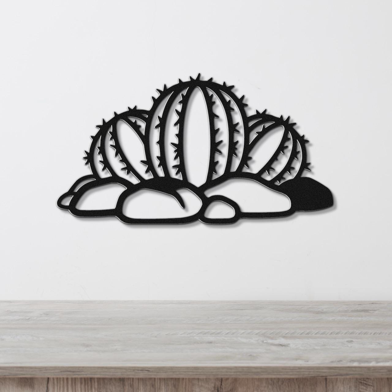 Black metal barrel cactus wall art with spines and cutout details, mounted on a white wall above a wooden surface.