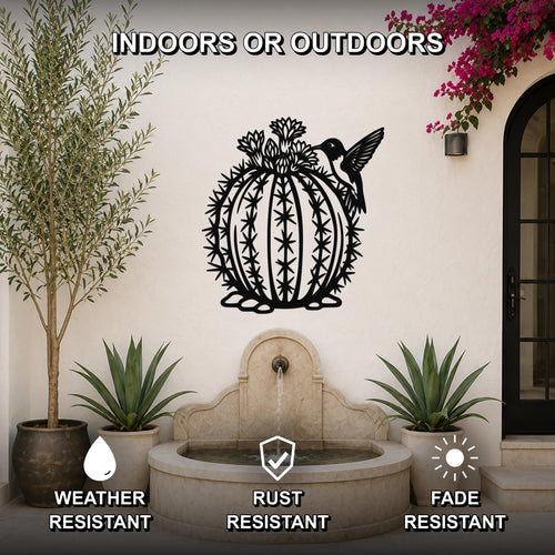 Black metal barrel cactus wall sculpture with flower and hummingbird cutouts, mounted on a white outdoor wall above a stone fountain.