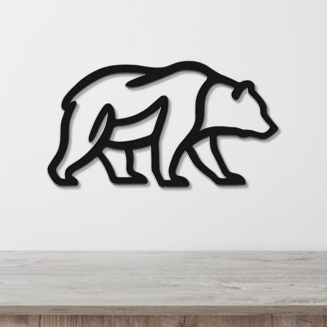 Black metal bear silhouette wall art with internal cutout details, mounted on a white wall above a light wood surface.