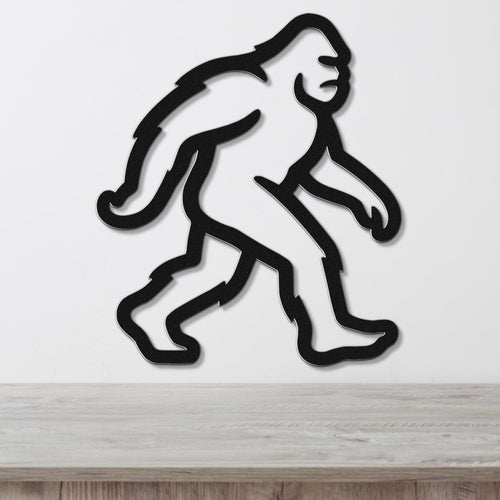 Black metal wall art cutout of a Bigfoot figure in a walking pose, mounted on a white wall above a wooden surface.