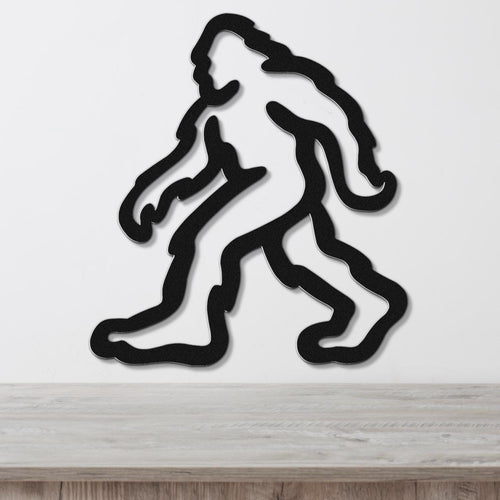 Black metal wall art featuring a minimalist Bigfoot silhouette with internal cutout details, mounted on a white wall above a light wood surface.