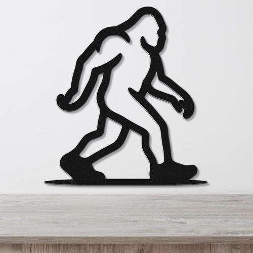 Black metal wall art silhouette of Bigfoot in a walking pose, mounted on a white wall above a light wood surface.