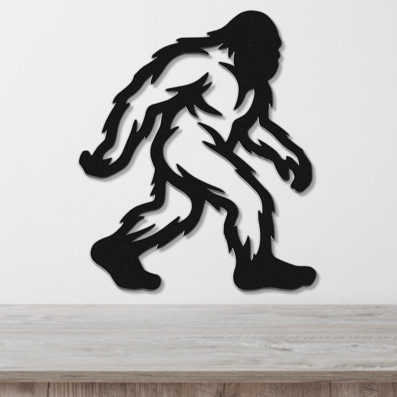 Black metal Bigfoot wall art silhouette with internal cutout details, mounted on a white wall above a wooden surface.