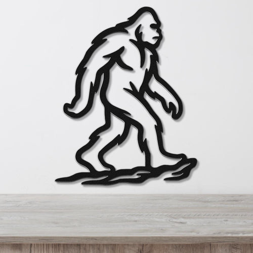 Black metal Bigfoot silhouette wall art with internal cutout details, mounted on a white wall above a light wood surface.