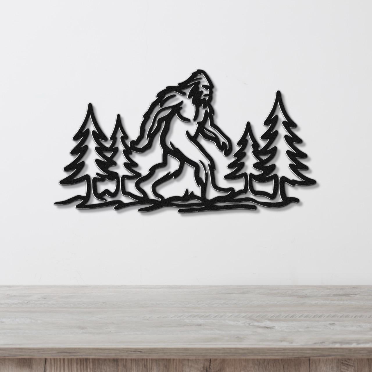Black metal wall art featuring a Bigfoot silhouette walking through pine trees, mounted on a white wall above a wooden counter.