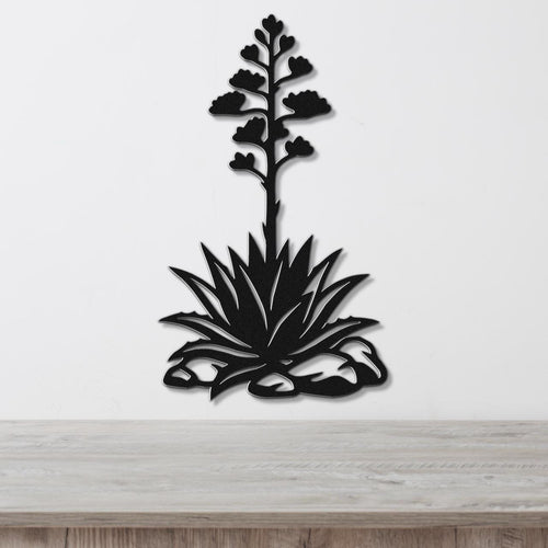 Black metal wall art silhouette of a blooming agave plant with a tall flower stalk and spiky leaves, mounted on a white wall above a wooden surface.