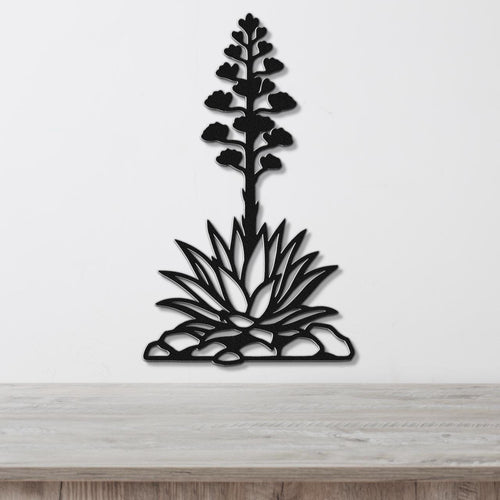 Black metal wall art in the silhouette of a blooming agave plant with cutout leaves and flower stalk, mounted on a white wall above a wooden surface.